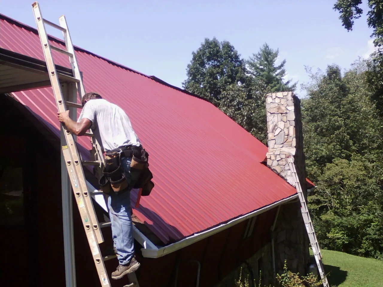 Roofer on a ladder beside a red metal roof and stone chimney — Soffit & Fascia Repair by Roofing Brothers in Monetta